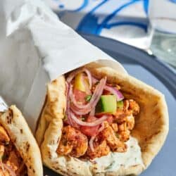 The doner kebab recipe with the cucumber tomato salad and tahini sauce on pita wrapped in a piece of parchment paper on a plate.