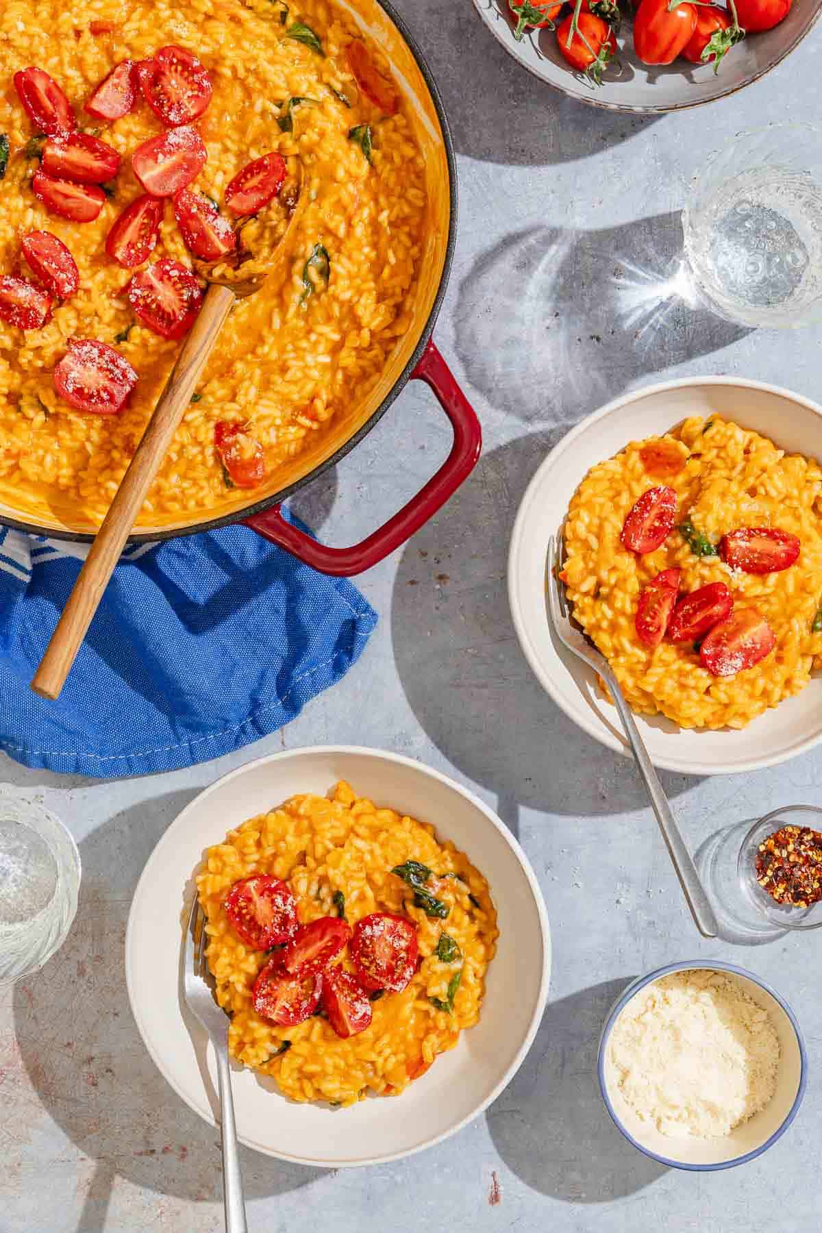 Two bowls of tomato risotto with forks next to bowls of Parmesan cheese, red pepper flakes and tomatoes, a glass of water, and the rest of the risotto in a pan with a wooden spoon.
