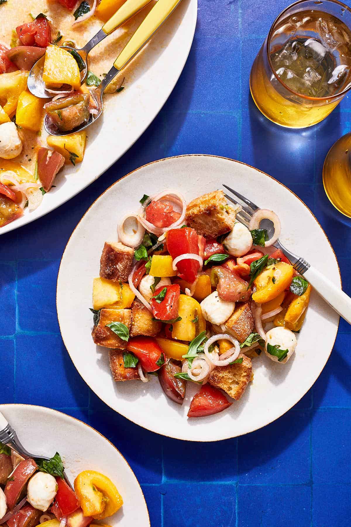 A serving of panzanella salad on a plate with a fork. Next to this is another plate of the salad, the rest of the salad on a serving platter and a glass of water.