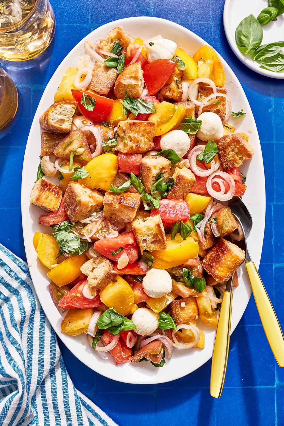 Panzanella salad on a serving platter with serving utensils. Next to this is a glass of ice water, a plate of fresh basil and a cloth napkin.