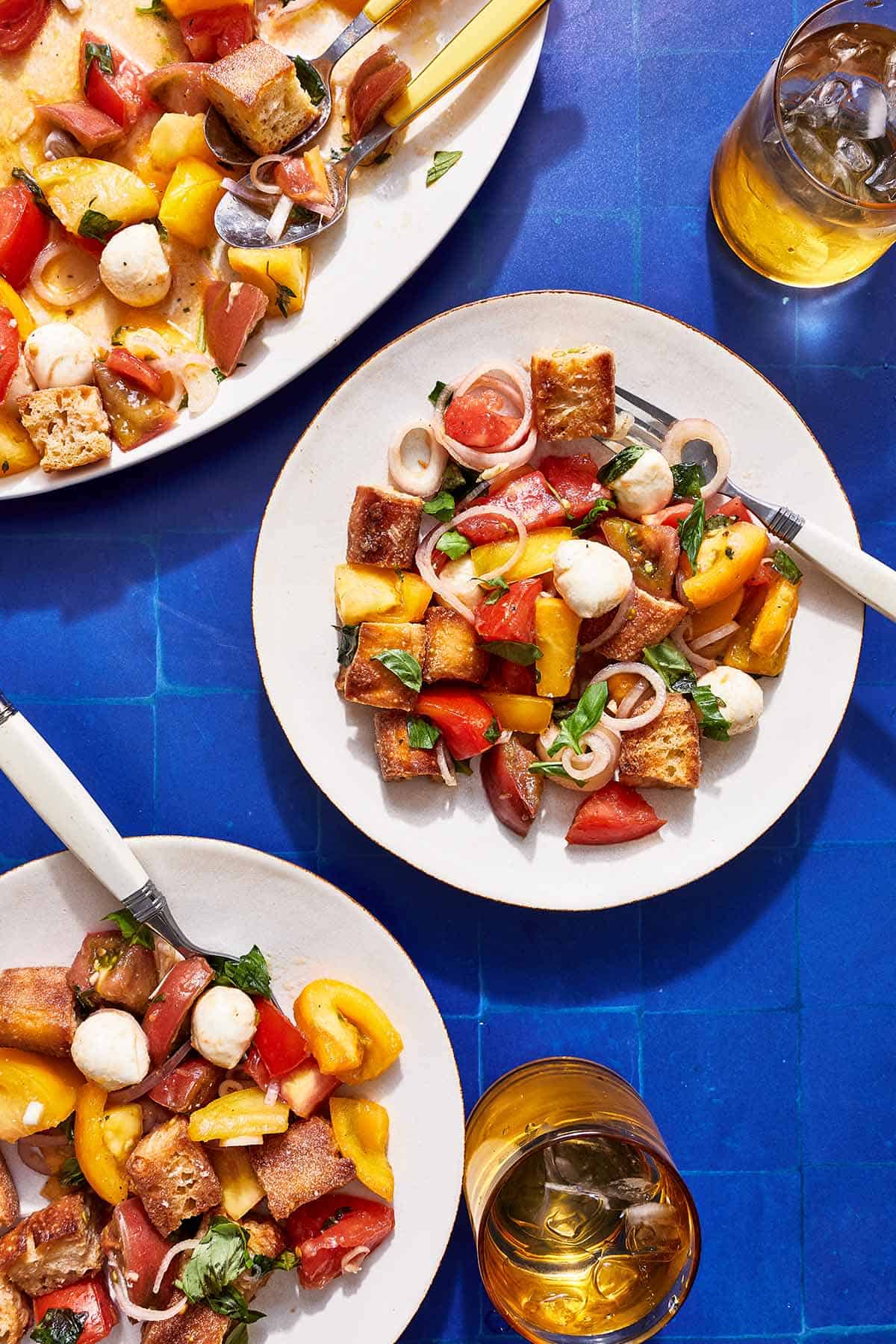A serving of panzanella salad on a plate with a fork. Next to this is another plate of the salad, the rest of the salad on a serving platter and two glasses of water.