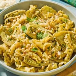 A serving of the artichoke pasta in a bowl next to a fork and a bowl of grated parmesan cheese.