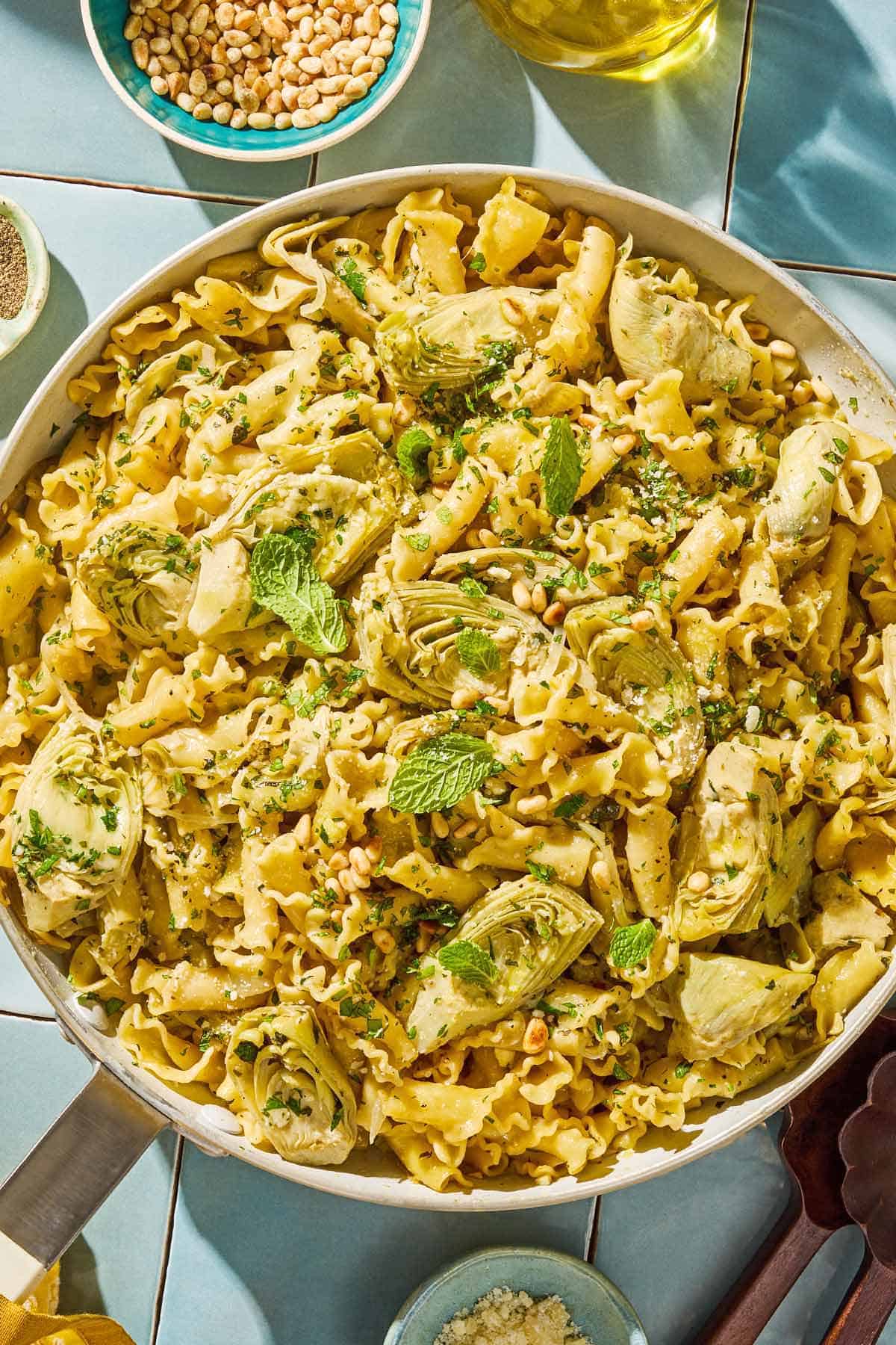 Artichoke pasta in a skillet topped with parmesan, pine nuts and mint. Next to this are bowls of grated parmesan cheese and pine nuts.
