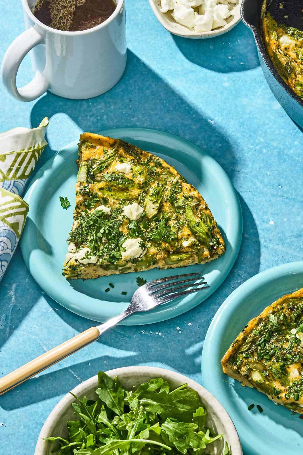 A slice of the asparagus frittata on a plate with a fork. Next to this is a cup of coffee, a bowl of goat cheese, another plate with a slice of the frittata, and a bowl of fresh herbs.