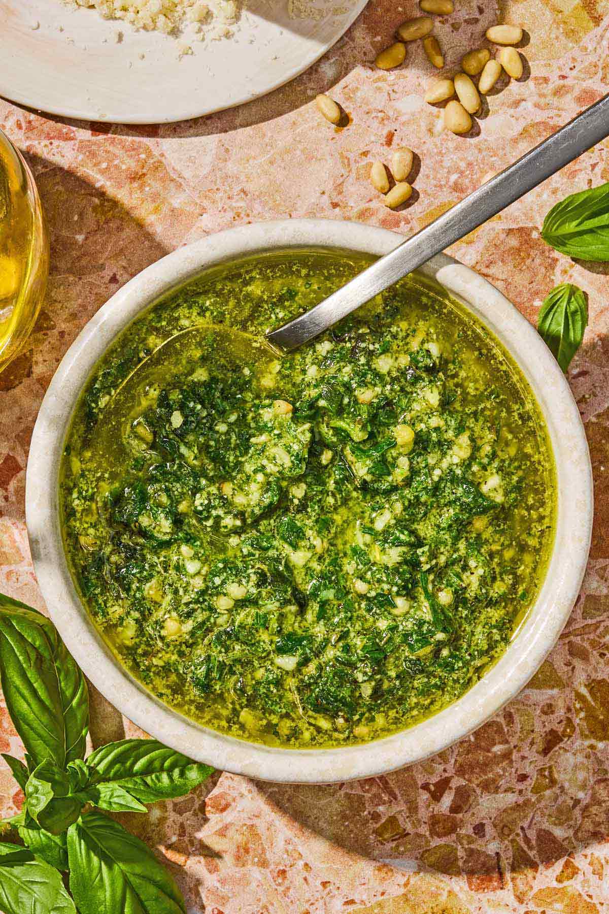 The basil pesto recipe in a bowl with a spoon surrounded by basil leaves and pine nuts.