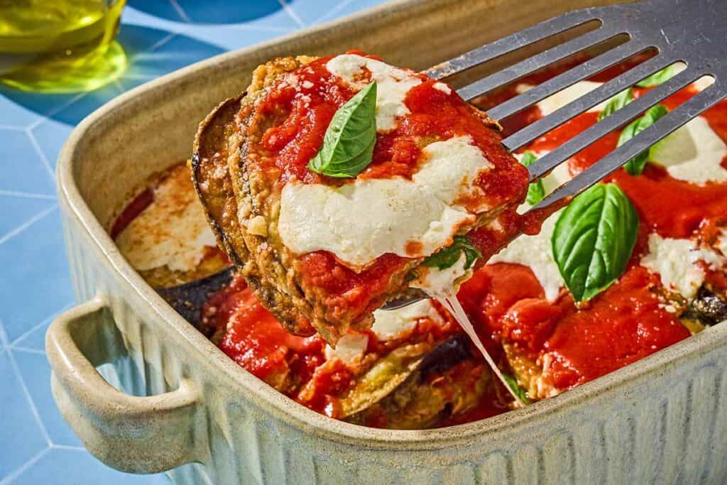 A piece of the eggplant parmesan being lifted from the rest of the casserole in a baking dish.