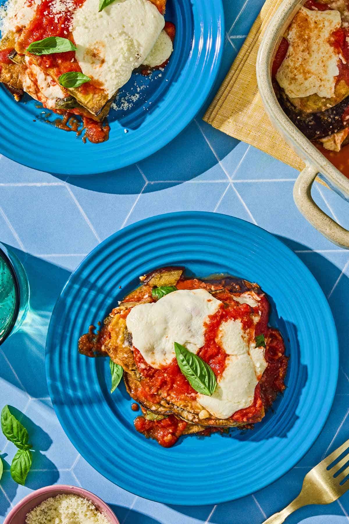 Two servings of the eggplant parmesan recipe on plates next to a fork and the rest of the eggplant parmesan in a baking dish.