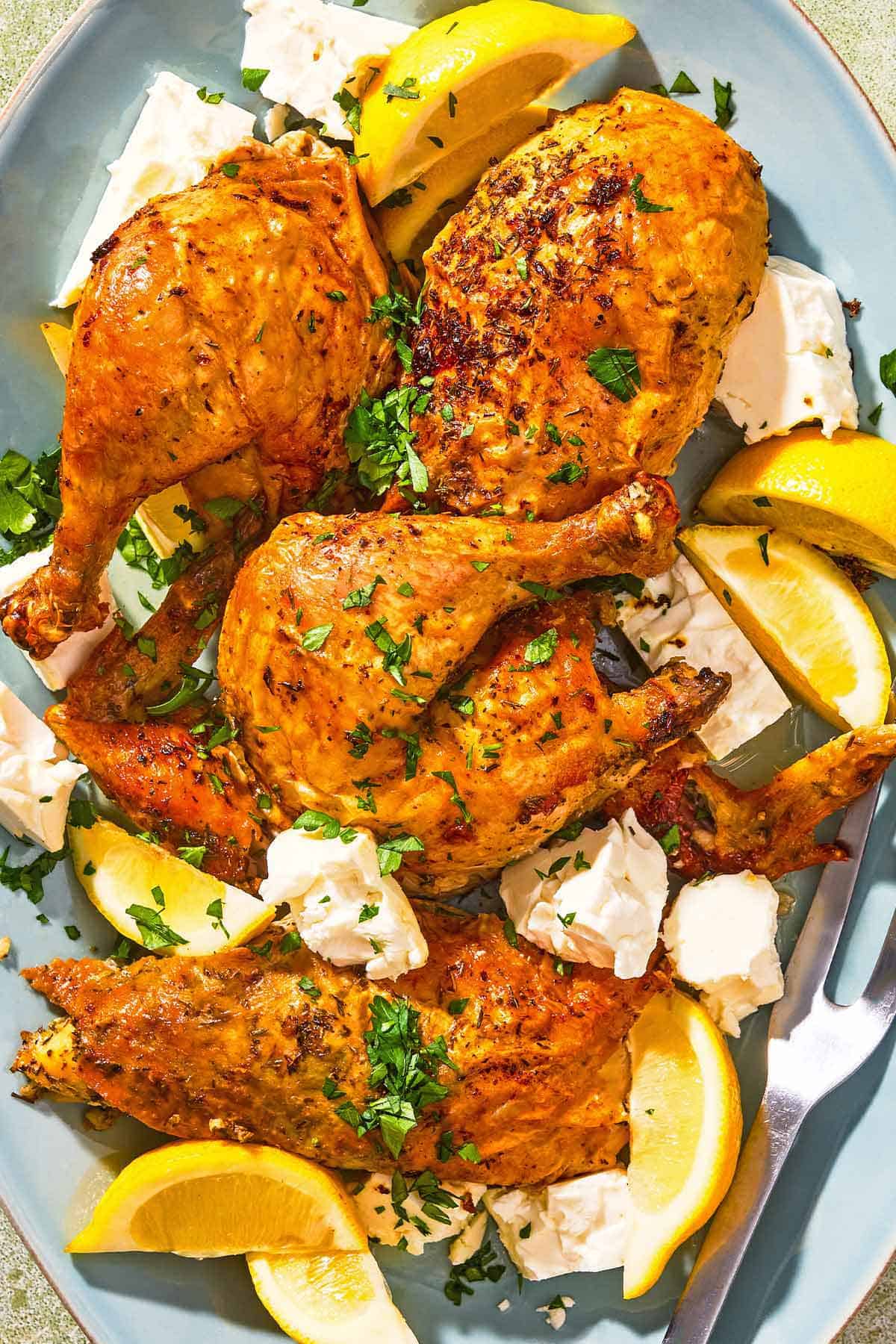 Feta-brined chicken on a platter with feta, lemon wedges and a serving fork.