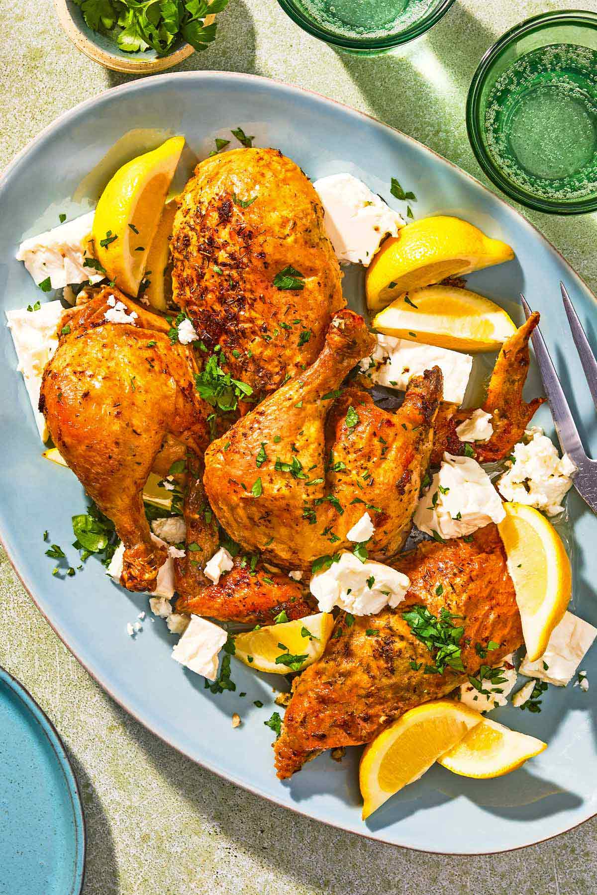 Feta-brined chicken on a platter with feta, lemon wedges and a serving fork. Next to this is a bowl of parsley, 2 glasses of sparkling water, and a plate.