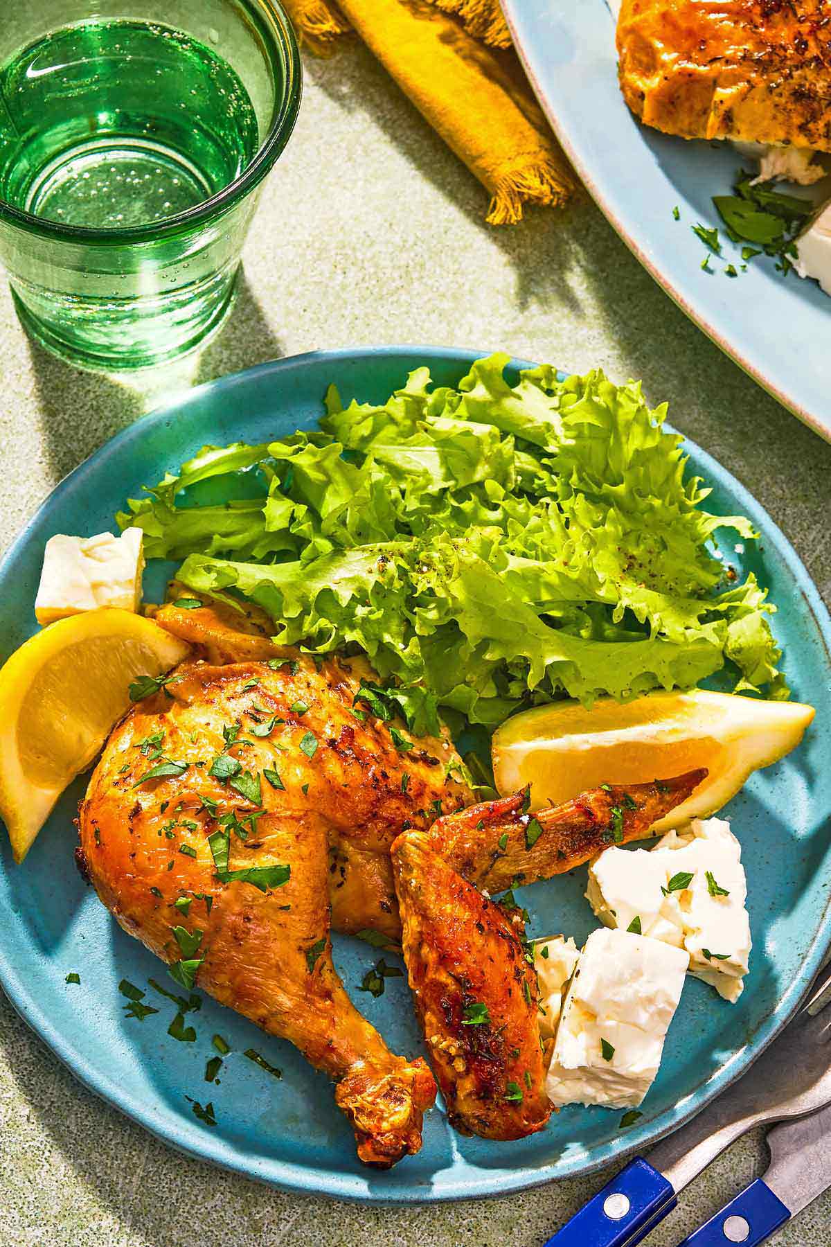 A serving of the feta brined chicken on a plate with feta cheese, lemon wedges and a salad next to a glass of water.