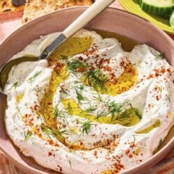 Haydari topped with fresh dill, Aleppo pepper and a drizzle of olive oil in a bowl with a spoon. Next to this is a bowl of sliced cucumbers and pita wedges.
