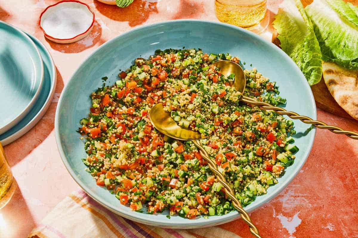 Quinoa tabouli in a serving bowl with serving utensils next to leaves of romaine, pita bread, a glass of sparkling water, a bowl of salt and a stack of 2 plates.