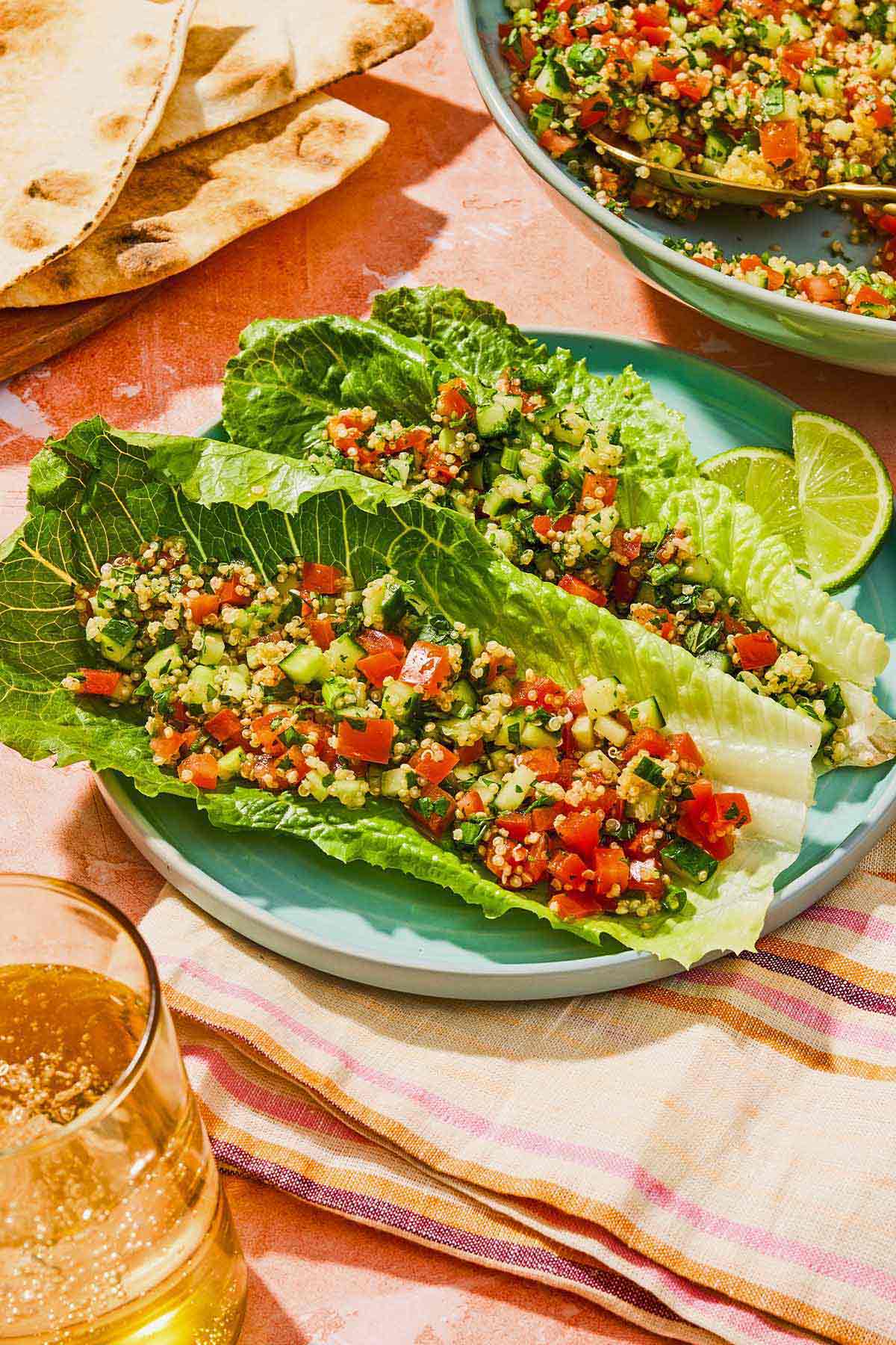Two romaine lettuce leaves topped with quinoa tabouli on a plate with lime wedges. Next to this is a glass of sparkling water, a stack of pita, and the rest of the quinoa tabouli in a serving bowl.