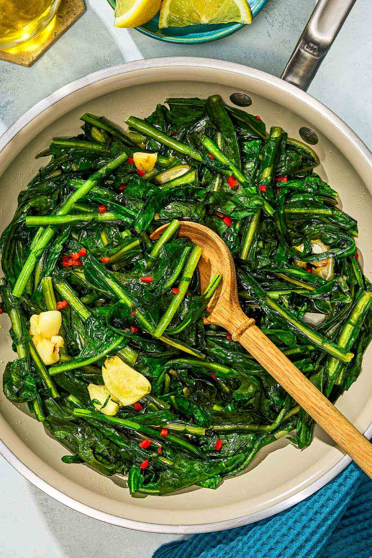 Dandelion greens in a skillet with a wooden spoon next to a bowl of lemon wedges.