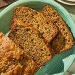 A partially sliced loaf of zucchini bread on a platter. Next to this is a stack of 2 plates with forks and a bowl of butter.
