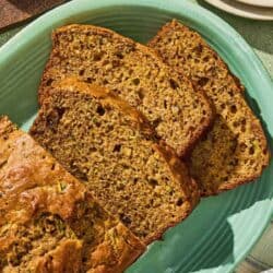 A partially sliced loaf of zucchini bread on a platter. Next to this is a stack of 2 plates with forks and a bowl of butter.