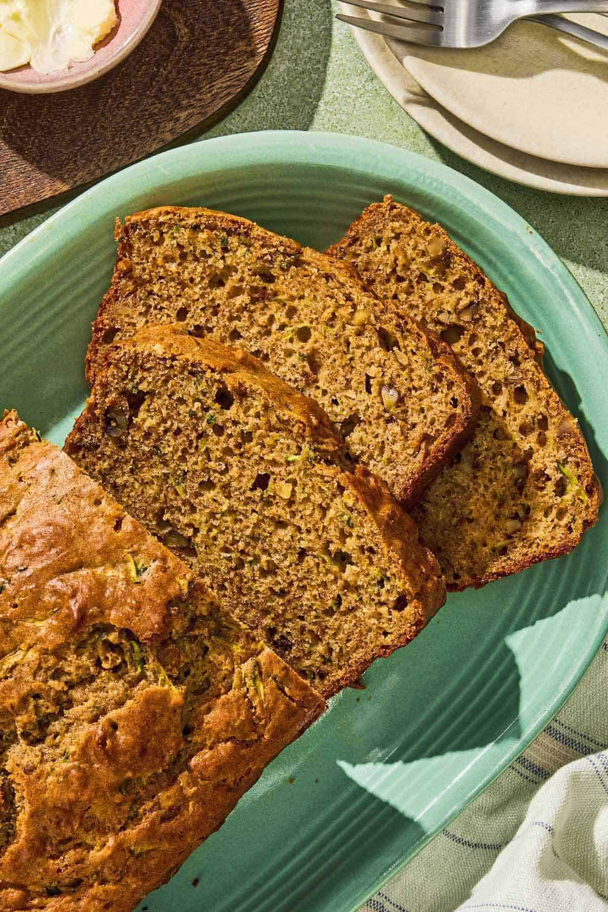 A partially sliced loaf of zucchini bread on a platter. Next to this is a stack of 2 plates with forks and a bowl of butter.
