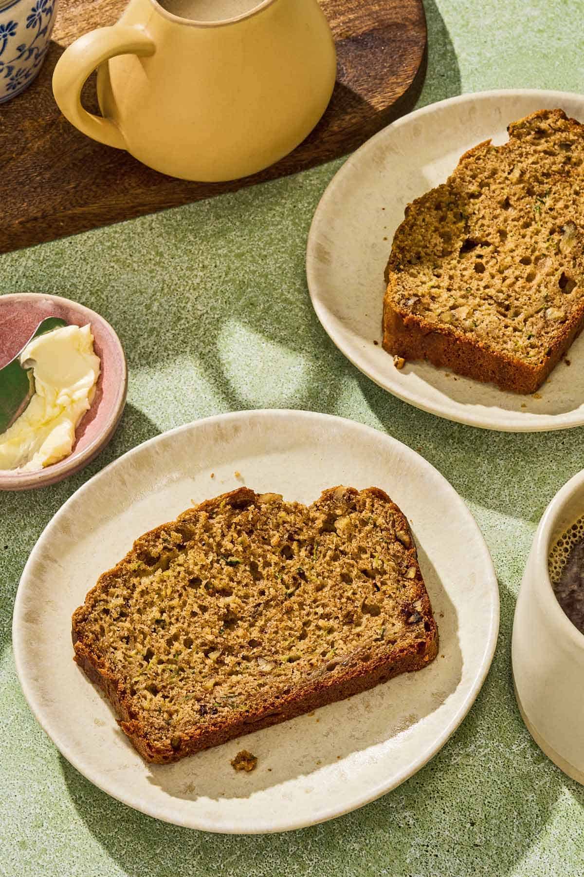 Two slices of zucchini bread on 2 plates next to a cup of coffee, a bowl of butter with a knife and a carafe of cream.