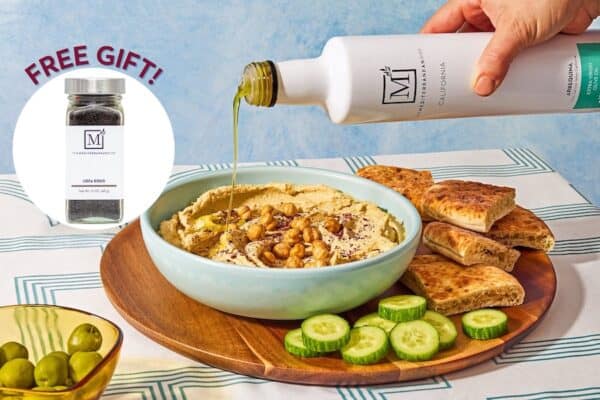 A bowl of hummus on a tray with pita and cucumbers with a drizzle of The Mediterranean Dish olive oil.