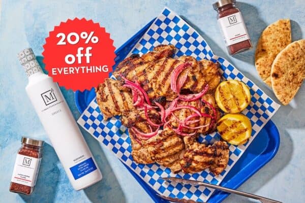 Grilled chicken on a platter with bottles of The Mediterranean Dish olive oil and spices with a 20% off promotional badge.