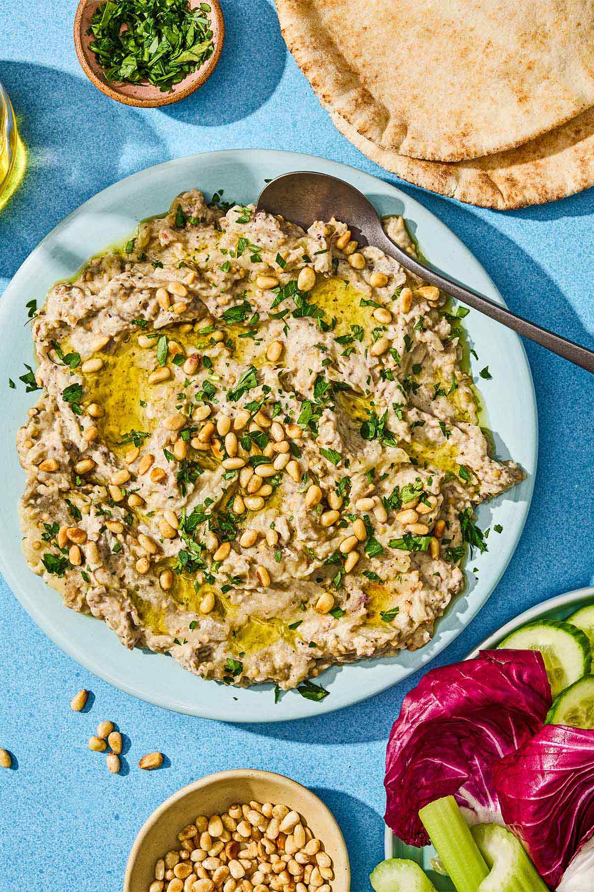 Baba ganoush in a bowl with a spoon next to a bowl of pine nuts, a bowl of fresh herbs, pita bread, and a plate of vegetables.