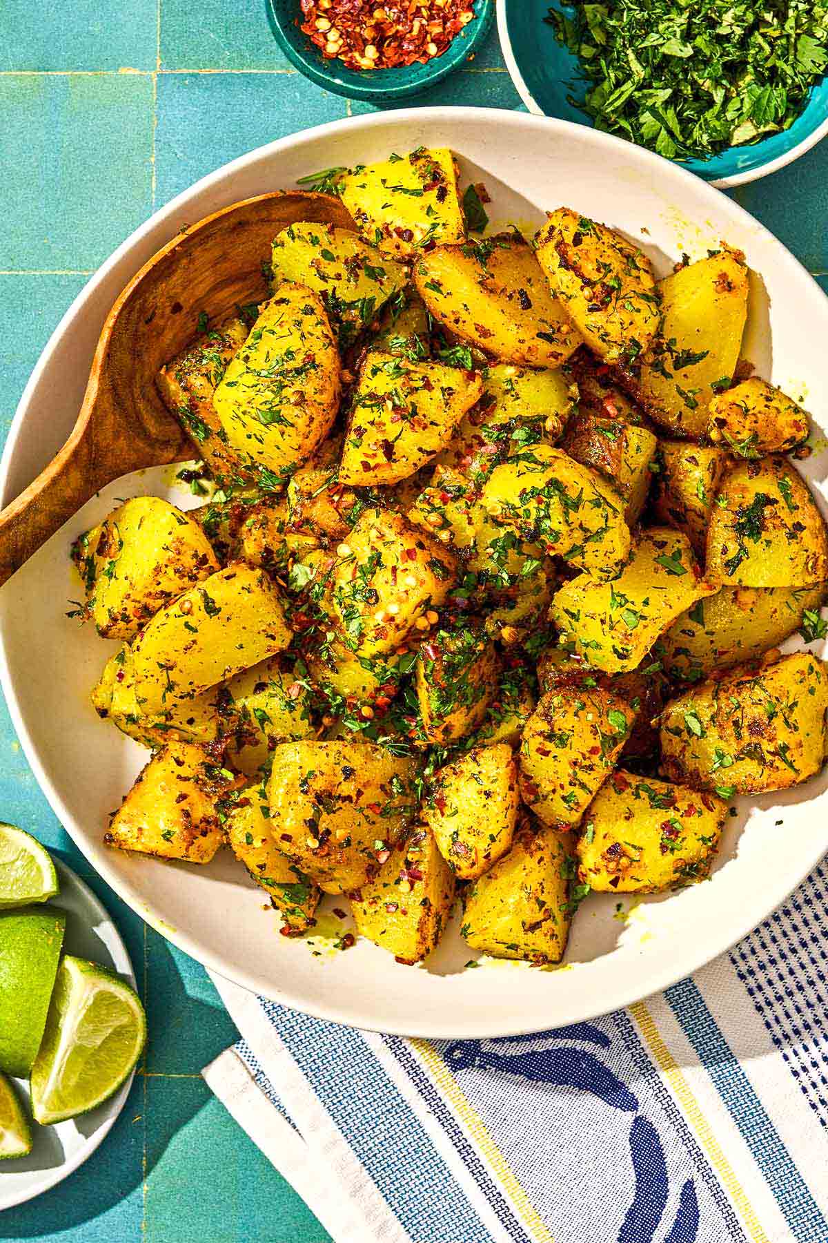 Batata Harra in a serving bowl with a spoon. Next to this is a plate with lime wedges and bowls of red pepper flakes and fresh herbs.