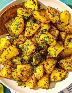 Batata Harra in a serving bowl with a spoon. Next to this is a plate with lime wedges and bowls of red pepper flakes and fresh herbs.
