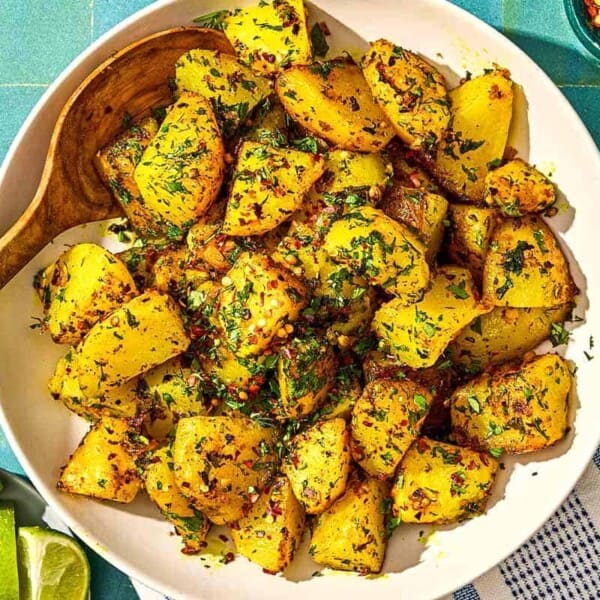 Batata Harra in a serving bowl with a spoon. Next to this is a plate with lime wedges and bowls of red pepper flakes and fresh herbs.