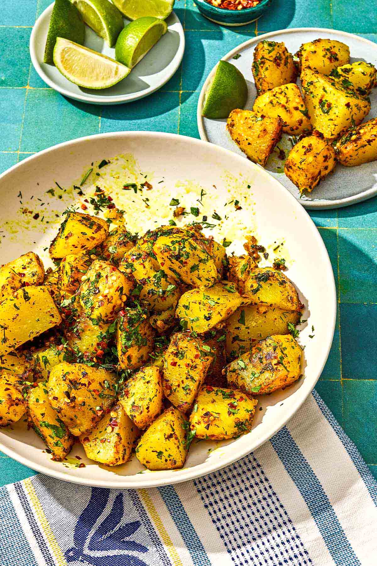 Batata Harra dans un bol de service avec une cuillère à côté d'une portion de la recette sur une assiette avec un quartier de citron vert et une assiette de quartiers de citron vert supplémentaires.