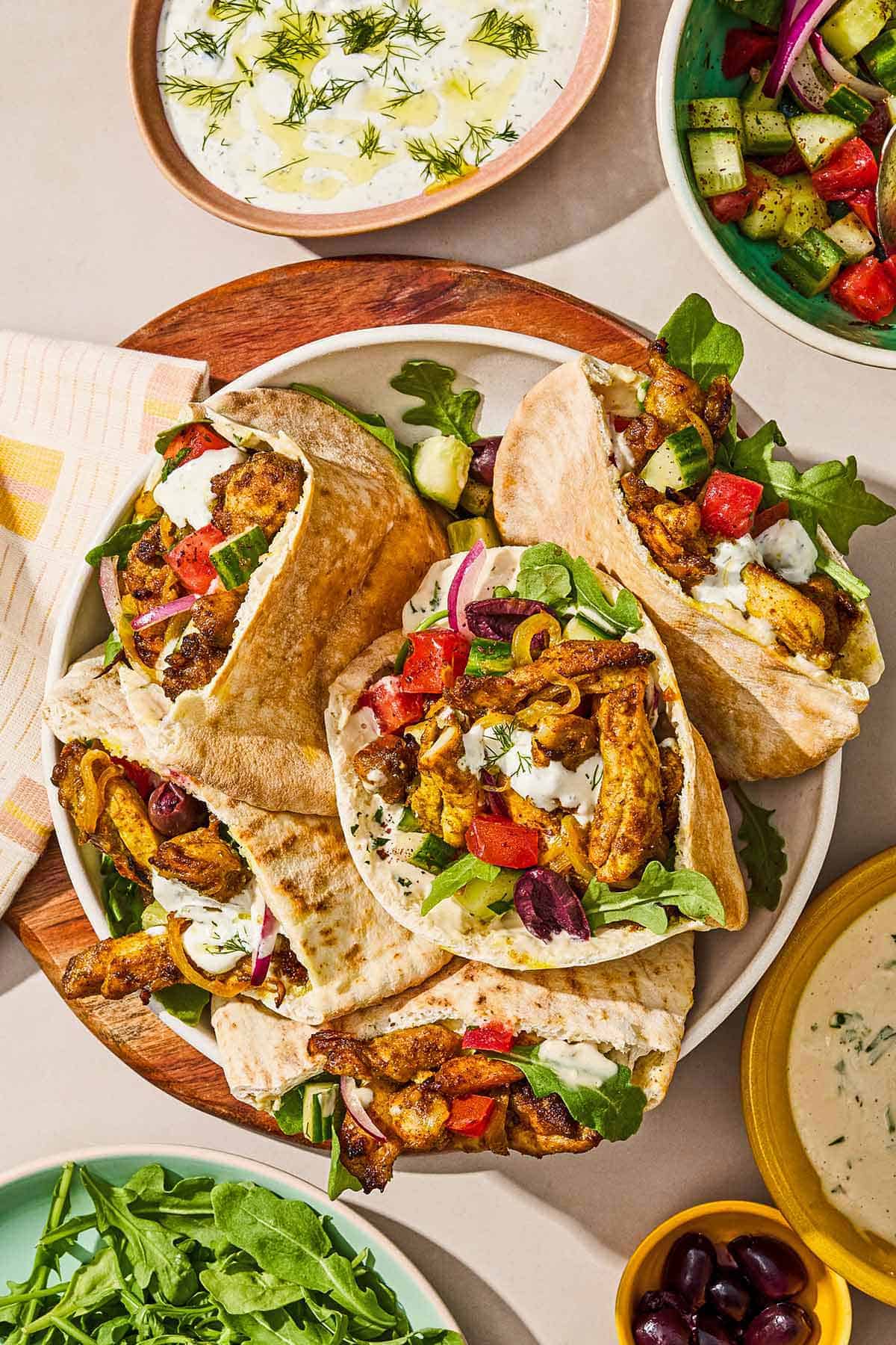 Several chicken shawarma pita sandwiches in a serving bowl on a wooden tray. Next to this are bowls of tahini sauce, tzaziki sauce, tomato cucumber salad and a plate of arugula.