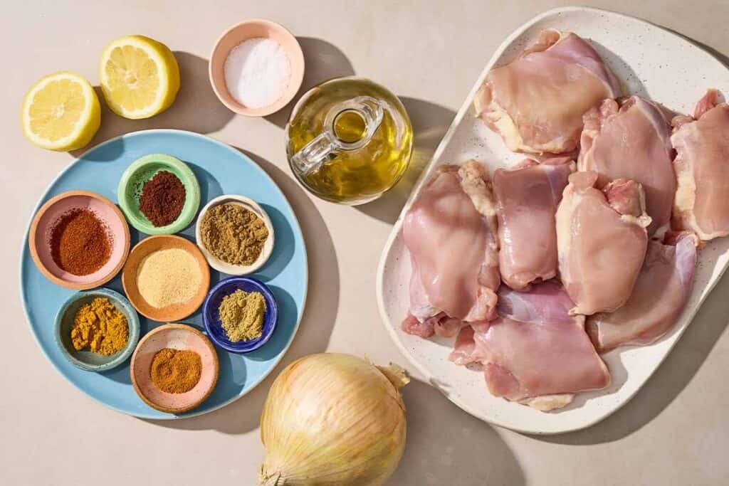 Ingredients for chicken shawama including chicken thighs, a yellow onion, olive oil, salt, lemon, cumin, turmeric, coriander, garlic powder, paprika, ground cloves, and cayenne pepper.