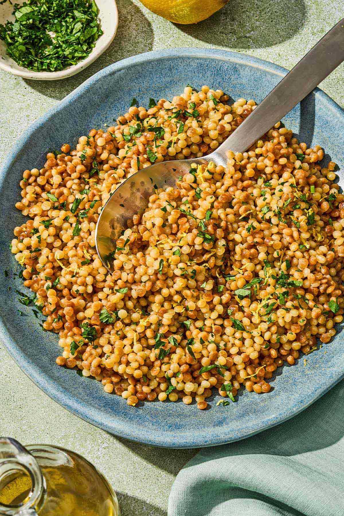 The pearl couscous recipe in a bowl with a spoon next to a bottle of olive oil and a bowl of chopped parsley.
