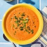 Gazpacho in a bowl next to a spoon, kitchen towel and a bottle of olive oil.