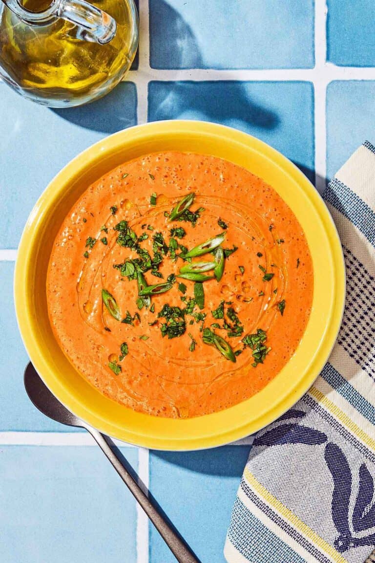 Gazpacho in a bowl next to a spoon, kitchen towel and a bottle of olive oil.