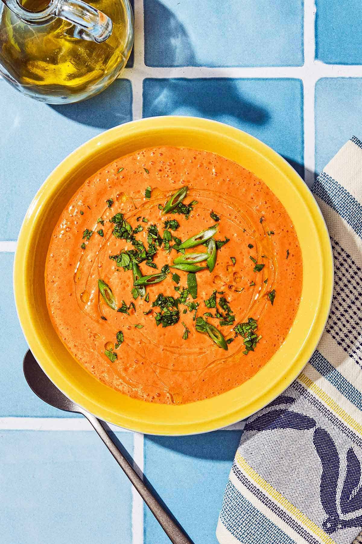 Gazpacho in a bowl next to a spoon, kitchen towel and a bottle of olive oil.