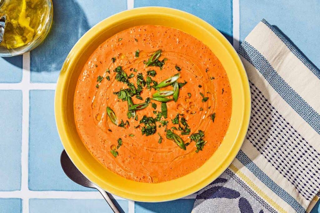 Gazpacho in a bowl next to a spoon, kitchen towel and a bottle of olive oil.