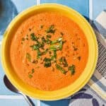 Gazpacho in a bowl next to a spoon, kitchen towel and a bottle of olive oil.