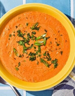Gazpacho in a bowl next to a spoon, kitchen towel and a bottle of olive oil.