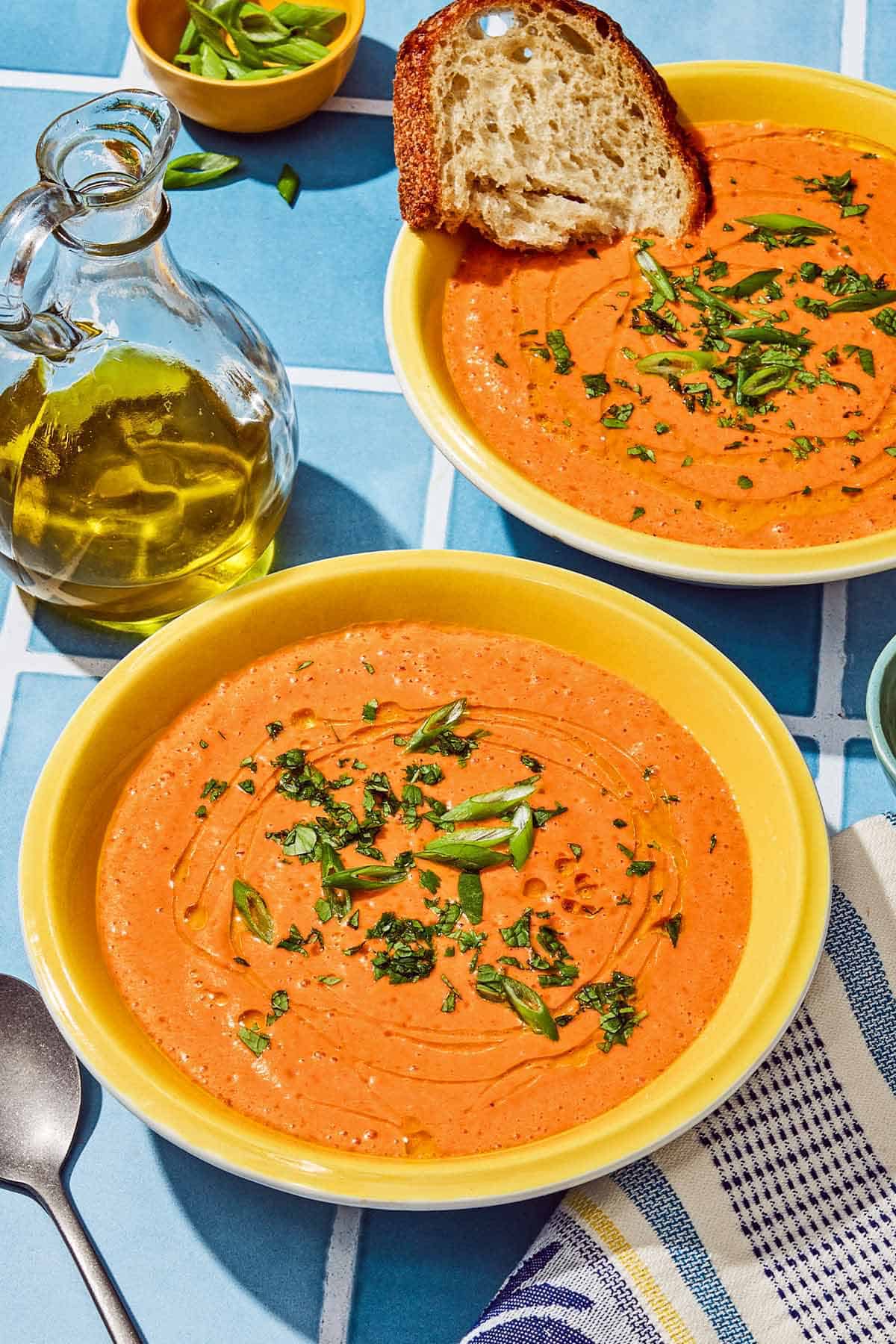 Two bowls of gazpacho, one with a slice of bread. Next to these is a spoon, a bottle of olive oil, a bowl of sliced green onions and a kitchen towel.