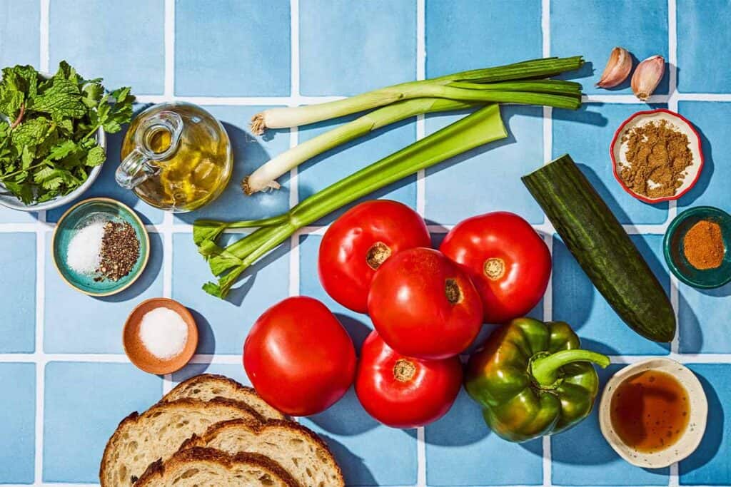 Ingredients for gazpacho including tomatoes, slices of bread, cucumber, celery, green pepper, green onions. garlic, olive oil, sherry vinegar, salt, pepper, cumin, cayenne, sugar, mint and cilantro.