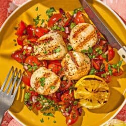 A serving of grilled scallops with Mediterranean salsa, a lemon half, a knife and a fork on a plate.