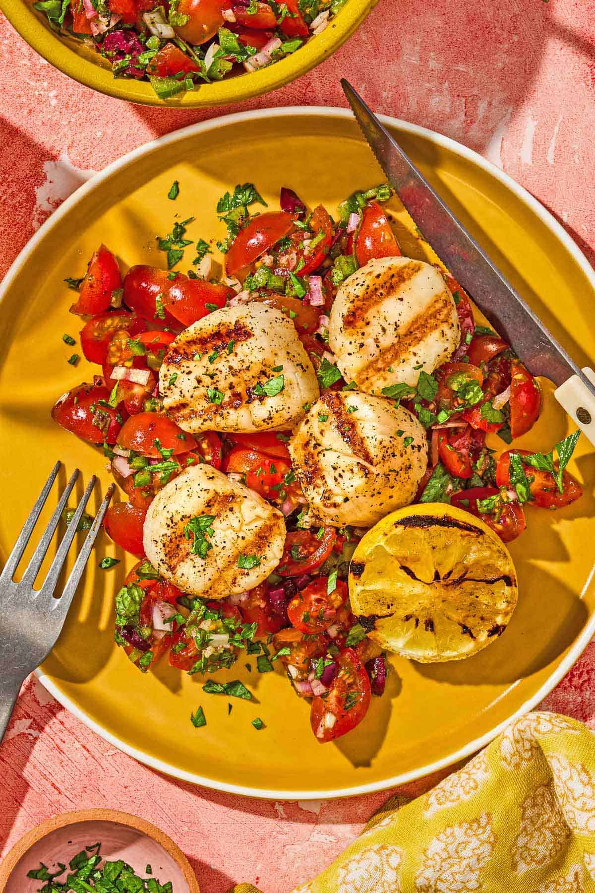 A serving of grilled scallops with Mediterranean salsa, a lemon half, a knife and a fork on a plate.