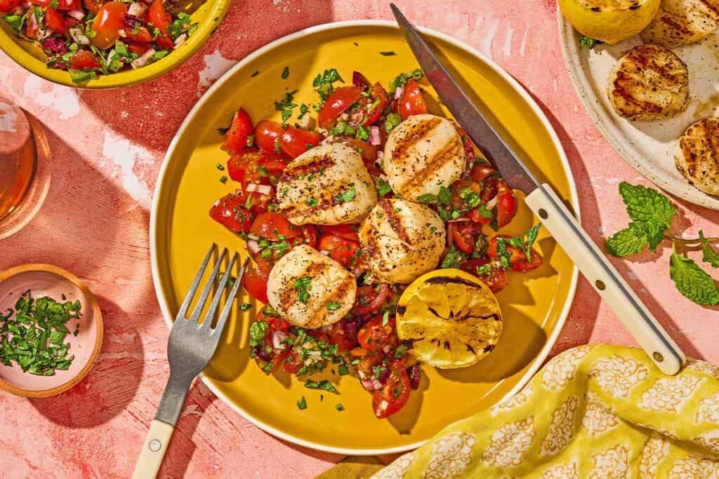 A serving of grilled scallops with Mediterranean salsa, a lemon half, a knife and a fork on a plate. Next to this is a bowl of chopped parsley, the Mediterranean salsa in a bowl, and the rest of the scallops on a plate.
