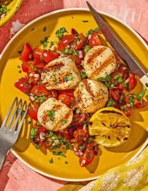 A serving of grilled scallops with Mediterranean salsa, a lemon half, a knife and a fork on a plate. Next to this is a bowl of chopped parsley, the Mediterranean salsa in a bowl, and the rest of the scallops on a plate.