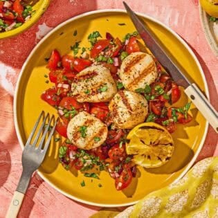 A serving of grilled scallops with Mediterranean salsa, a lemon half, a knife and a fork on a plate. Next to this is a bowl of chopped parsley, the Mediterranean salsa in a bowl, and the rest of the scallops on a plate.