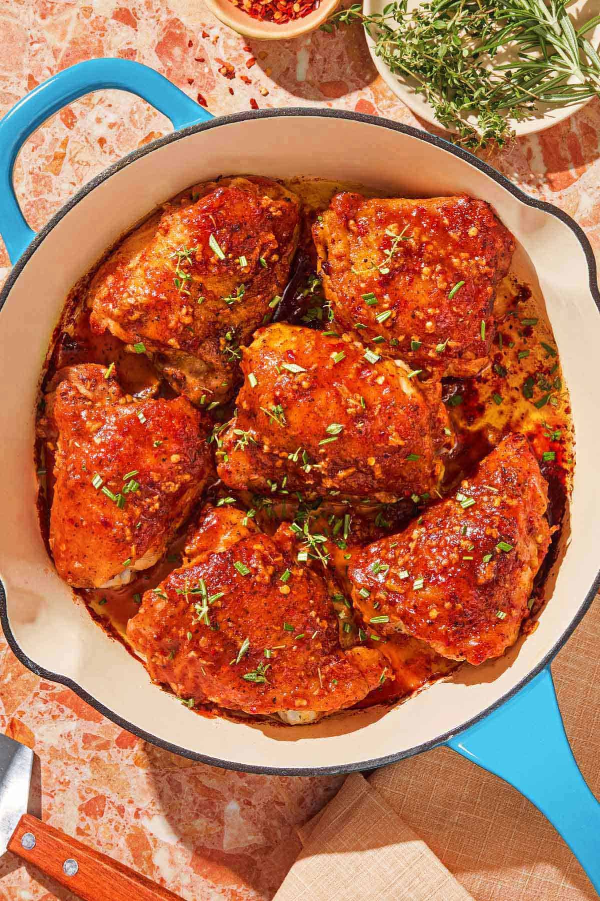 Honey mustard chicken in a skillet next to a bowl of red pepper flakes and a plate of fresh herbs.