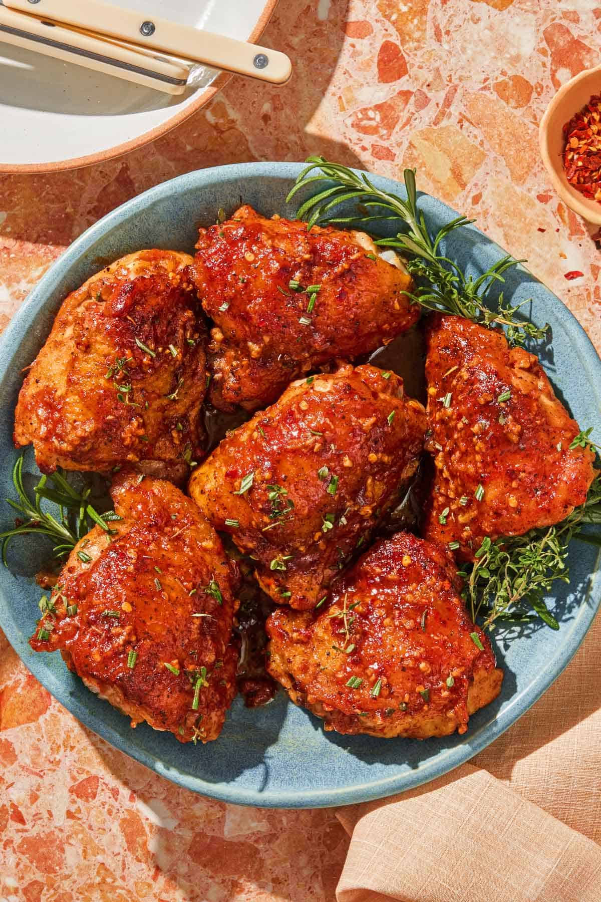 Honey mustard chicken on a platter with fresh herbs. Next to this is a bowl of red pepper flakes and a plate with utensils.