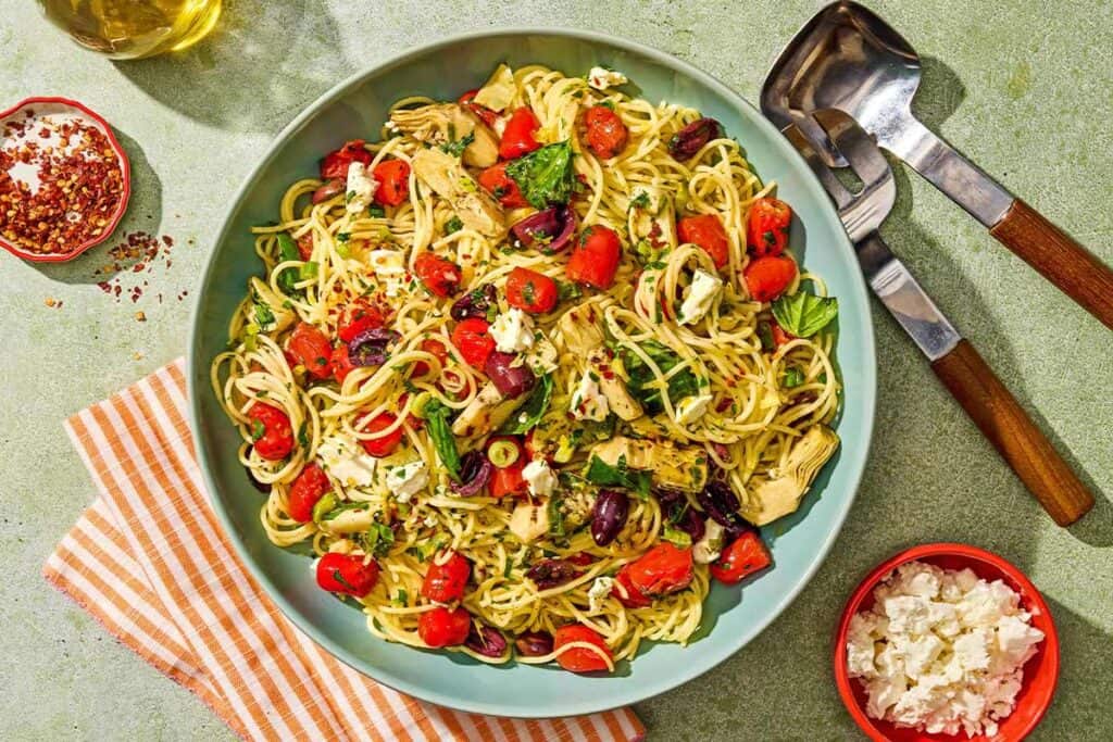 The Mediterranean pasta in a serving bowl next to serving utensils and bowls of feta and red pepper flakes.
