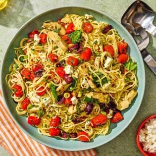 The Mediterranean pasta in a serving bowl next to serving utensils and bowls of feta and red pepper flakes.