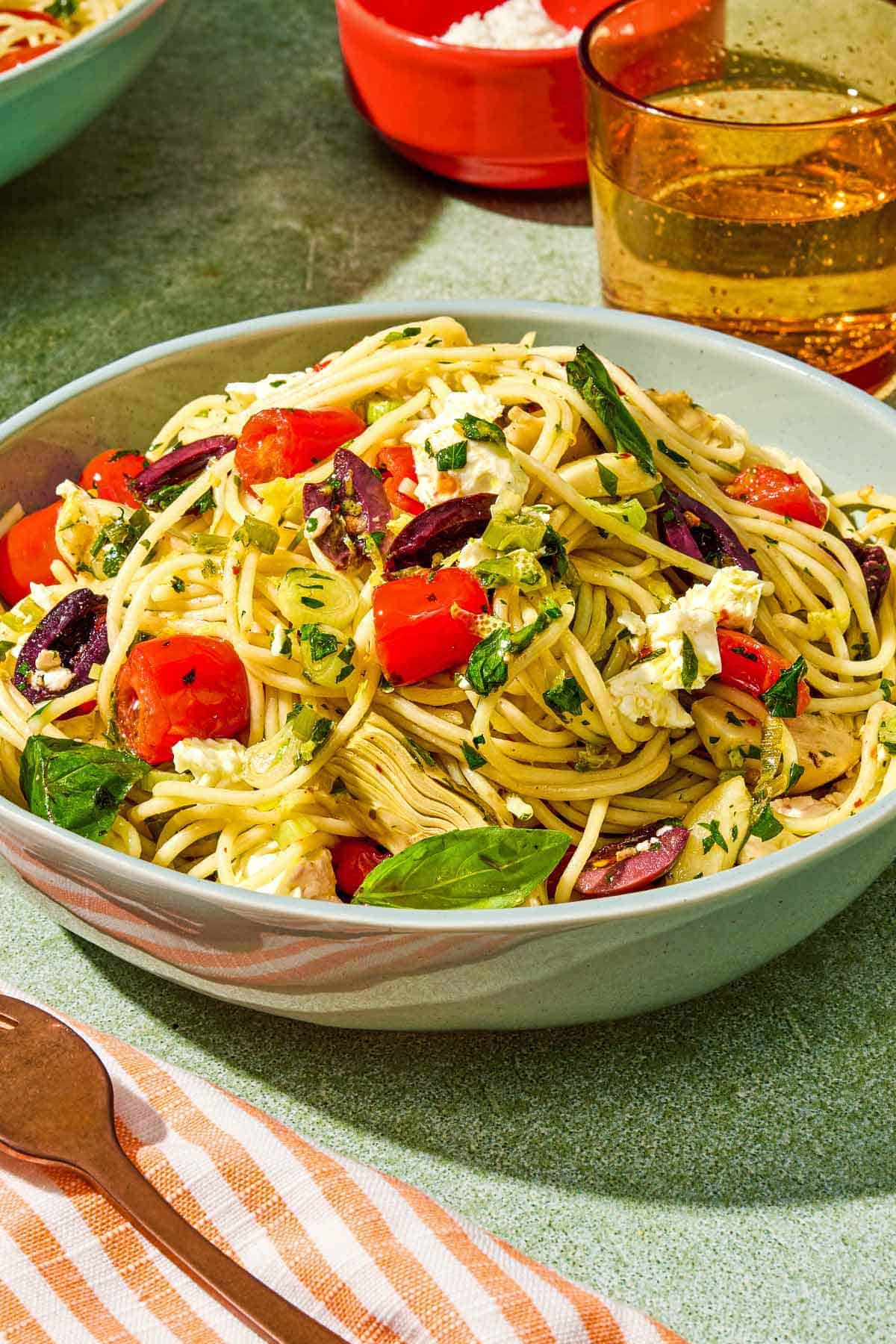 A serving of Mediterranean pasta in a bowl next to a fork, a bowl of feta and a glass of sparkling water.